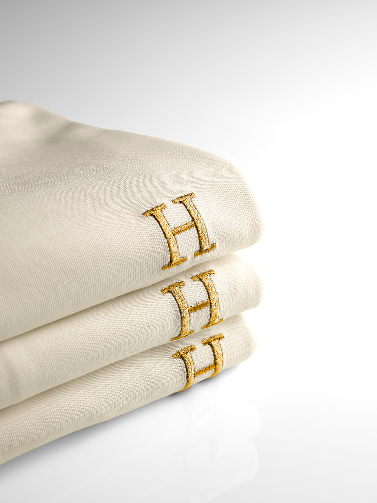 Drie creme HUMM Tees opgevouwen op elkaar, met op elk de goud geborduurde H