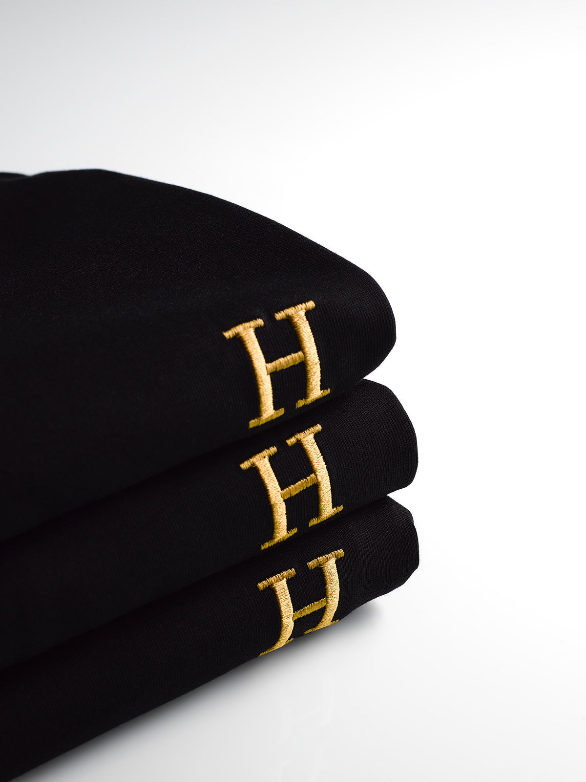 Drie zwarte HUMM Tees opgevouwen op elkaar, met op elk de goud geborduurde H