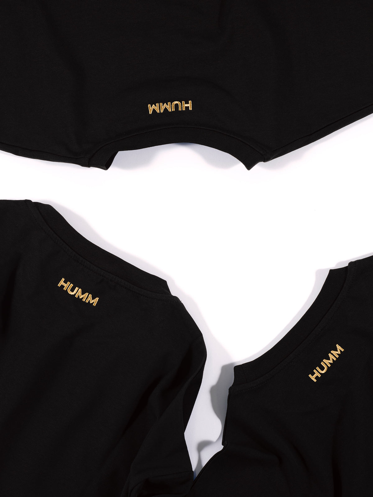 Drie zwarte HUMM Tees van bovenaf, met het goud geborduurde HUMM-woordmerk in de nek