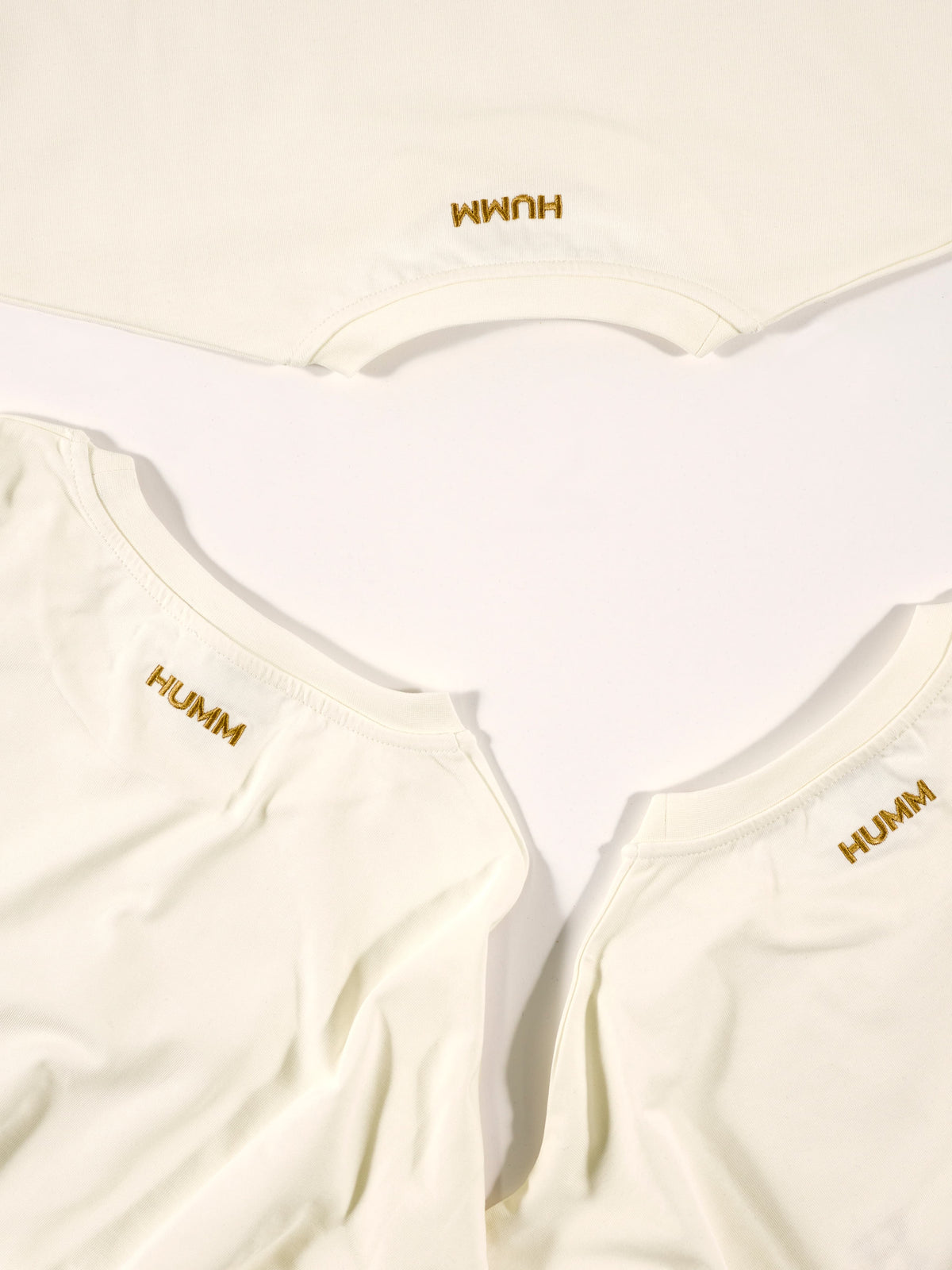Drie creme HUMM Tees van bovenaf, met het goud geborduurde HUMM-woordmerk in de nek